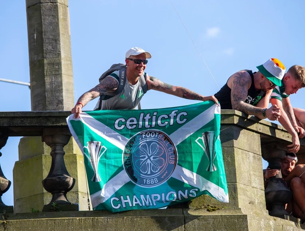 Ein treuer Anhänger zeigt stolz eine grüne Vereinsfahne mit der Aufschrift „Champions“ von einer Brücke. Erleben Sie den puren Erfolg und die Emotionen schottischer Traditionsvereine.